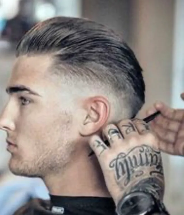A slick-back with a skin fade being finished, the barber's tattooed hands at work